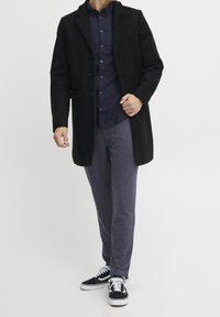 Schwarzer Wollmantel mit Reverskragen, dunkelblaues Hemd mit Knöpfen, graue Hose und schwarze Sneakers mit weißen Sohlen. Einfaches und modernes Design.