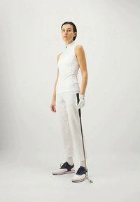 Λευκή αμάνικη μπλούζα με ψηλό γιακά, λευκό εφαρμοστό παντελόνι, ναυτικού μπλε και λευκά παπούτσια, κρατώντας ένα μπαστούνι του γκολφ. Λείο ύφασμα και καθαρές γραμμές.