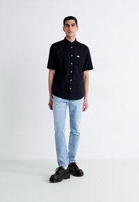 Camicia nera a maniche corte con bottoni e tasca sul petto, abbinata a jeans in denim blu chiaro e scarpe con piattaforma nera allacciate.