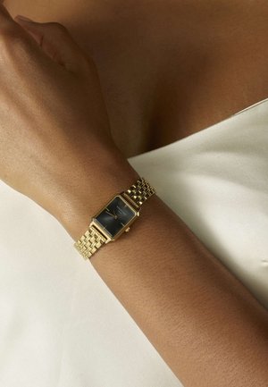Goldfarbene rechteckige Uhr mit einem schwarzen Ziffernblatt, strukturiertem goldenen Armband und dezenten Stundenmarkierungen. Am Handgelenk getragen, vor einem hellen Stoffhintergrund.