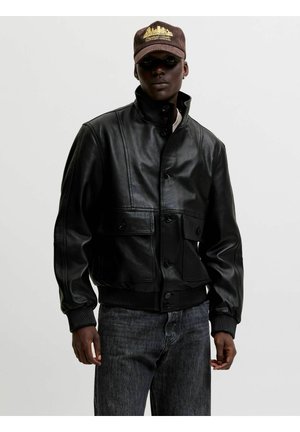 Osoba nosząca czarną skórzaną kurtkę zapinaną na guziki z kieszeniami z patkami, ciemne dżinsy oraz brązową czapkę z żółtym haftem panoramy miasta.