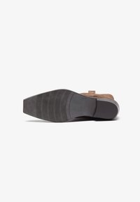 Zapato de ante marrón con punta cuadrada y taco bajo. La suela de goma negra presenta ranuras horizontales para mayor tracción. Diseño minimalista.