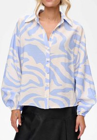 Vrouw in een losse blouse met een blauw-wit abstract patroon, lange pofmouwen, knoopsluiting aan de voorkant, en een zwarte leren rok.