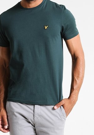 Donkergroen katoenen t-shirt met een ronde hals en een klein geel logo op de linkerborst. G gedragen met lichtgrijze broek.