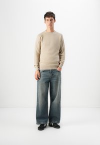 Giovane uomo che indossa un maglione beige, jeans a gamba larga blu sbiaditi e sneaker nere, in piedi con una mano in tasca su sfondo bianco.