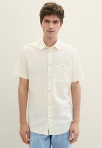 Cămașă cu mânecă scurtă, de culoare crem, cu butoane în față, buzunar frontal, textură subtilă și butoane mici, maro, purtată cu blugi din denim de culoare albastru deschis.