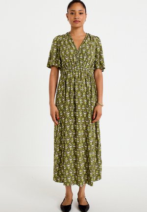 Vrouw draagt een groene midi-jurk met bloemenprint, korte mouwen en zwarte ballerina's, staand tegen een effen witte achtergrond.