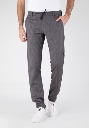 Mężczyzna w szarych spodniach w prążki z ściągaczem w talii i białych trampkach, stojący z jedną ręką w kieszeni.