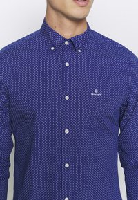 Camisa de botones azul marino con un pequeño patrón blanco. Fabricada con tela texturada, cuenta con un collar y un logotipo bordado en el pecho.