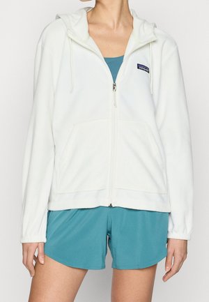 Woman wearing a white zip-up hooded fleece jacket over teal athletic tank top and matching shorts, standing with arms relaxed by sides.