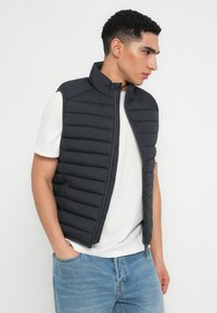 Sort puffervest med høj krave og quiltet design. Lynlås lukning og sidelommer. Bæres over en hvid skjorte og lys blå jeans.