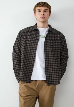 Schwarz-braun karierter Flanellhemd, zugeknöpft, über einem weißen Grafik-T-Shirt getragen; kombiniert mit beigen Hosen. Einfaches Design, auffällige Tasche.