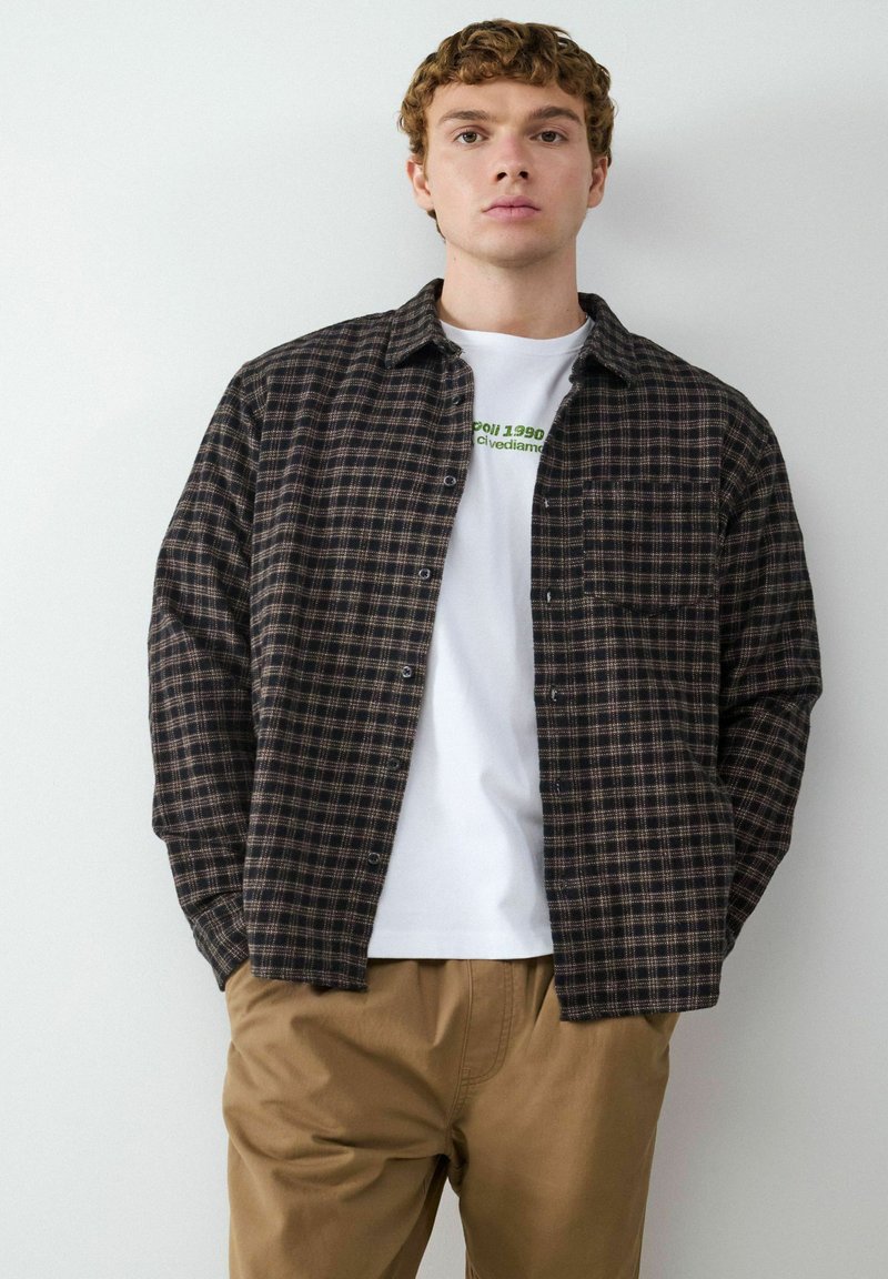 Zwarte en bruine geblokte flanellen shirt, geknoopt, gedragen over een wit T-shirt met een opdruk; gecombineerd met beige broek. Eenvoudig ontwerp, opvallende zak.