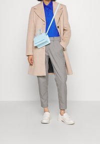 Manteau en laine beige, chemise bleu à boutons, pantalon à carreaux gris, chaussures blanches à plateforme, et un petit sac à main bleu clair avec une courte anse.