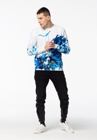 Camisa de manga larga en blanco con un patrón abstracto azul de agua, combinada con pantalones negros y zapatillas blancas. Tejido suave, corte relajado.