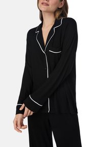Haut de pyjama noir à manches longues, avec des bordures blanches le long du col et de la poche, fermeture à boutons et coupe décontractée. Tissu au toucher doux.