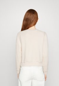 Sudadera beige de manga larga, con cuello redondo y dobladillo acanalado; textura suave con un diseño recortado. Modelo lleva pantalones blancos.