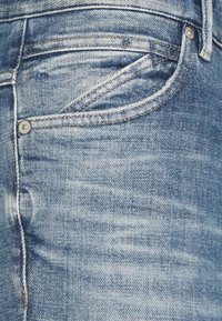 Närbild av bleka blå jeans som visar framfickan, myntfickan, bältet, och metallnitar med synliga sömnadsdetaljer.