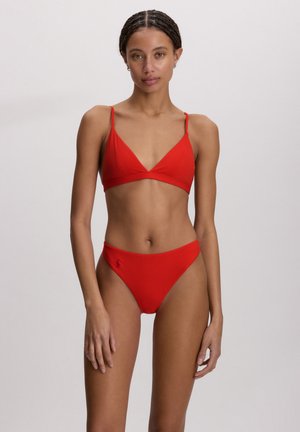 Γυναίκα με πλεξούδες που φοράει κόκκινο μαγιό δύο κομματιών, στέκεται μπροστά σε ένα απλό φωτεινό φόντο, κοιτάζοντας προς τα εμπρός.