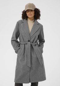 Manteau gris ceinturé en tissu au toucher lisse. Revers crantés, fermeture à un bouton et poches latérales. Porté sur un haut beige.