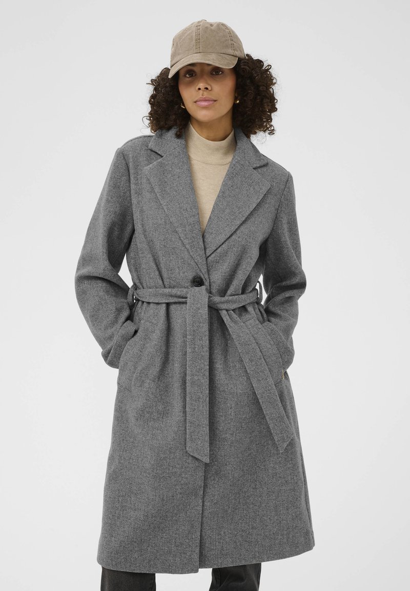 Manteau gris ceinturé en tissu au toucher lisse. Revers crantés, fermeture à un bouton et poches latérales. Porté sur un haut beige.