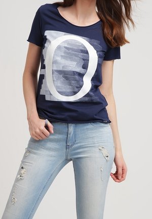 Frau trägt ein marineblaues T-Shirt mit einem großen weißen "O"-Grafik und hellblaue, zerrissene Skinny-Jeans, posiert mit einer Hand in der Tasche.