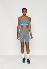 Τιρκουάζ αθλητικό σουτιέν με λογότυπο "UNDER ARMOUR", σε συνδυασμό με γκρι σορτς ψηλόμεσα. Το μοντέλο φοράει λευκές κάλτσες και μαύρα αθλητικά παπούτσια.