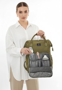 Olivgrön skötväska i ryggsäcksstil med vadderad textur, dubbla handtag på toppen och dragkedja. Inuti finns grå fack för flaskor och tillbehör.