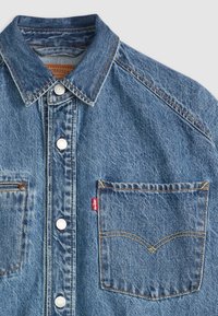 Lys blå denimskjorte med klassisk krage, knappelukking og en brystlomme med en særegen buet søm og rød etikett.