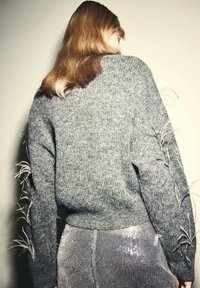Pull en tricot gris avec des manches longues et une broderie texturée. Le dos présente une coupe décontractée. Associé à un pantalon pailleté.