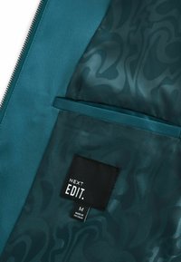 Teal kangas siseruumides, millel on paisley muster, must silt, millel on kirjas "NEXT EDIT". Suurus "M" ja nähtav tekst "MADE IN VIETNAM".
