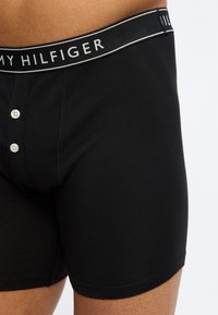 Κοντινό πλάνο ενός ατόμου που φοράει μαύρα μποξεράκια Tommy Hilfiger με δύο λευκά κουμπιά στο μπροστινό φερμουάρ και λωρίδα μέσης με λογότυπο.