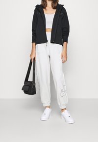 Svart zip-hoodie över en grå croptop, tillsammans med vita sweatpants med en Nike-logotyp och vita sneakers. Svart axelremsväska.