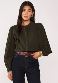 Camicia verde oliva a bottone con scollatura tonda e maniche a sbuffo. Abbinata a jeans scuri e una cintura trapuntata bordeaux. Accenti di gioielli dorati.
