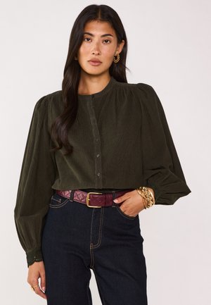 Camicia verde oliva a bottone con scollatura tonda e maniche a sbuffo. Abbinata a jeans scuri e una cintura trapuntata bordeaux. Accenti di gioielli dorati.