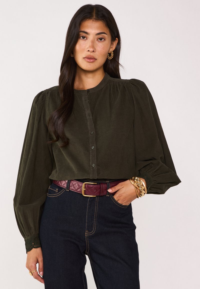 Camicia verde oliva a bottone con scollatura tonda e maniche a sbuffo. Abbinata a jeans scuri e una cintura trapuntata bordeaux. Accenti di gioielli dorati.