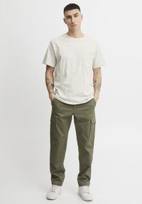 Beige színű pamut, kerek nyakú póló elülső zsebbel, olajzöld cargo nadrággal párosítva, ami oldalsó zsebekkel rendelkezik, valamint fehér sportcipővel.
