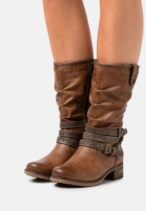Bottes montantes en faux cuir marron avec tige ample, sangles décoratives, clous et fermeture éclair latérale. Talon bas épais pour plus de stabilité.