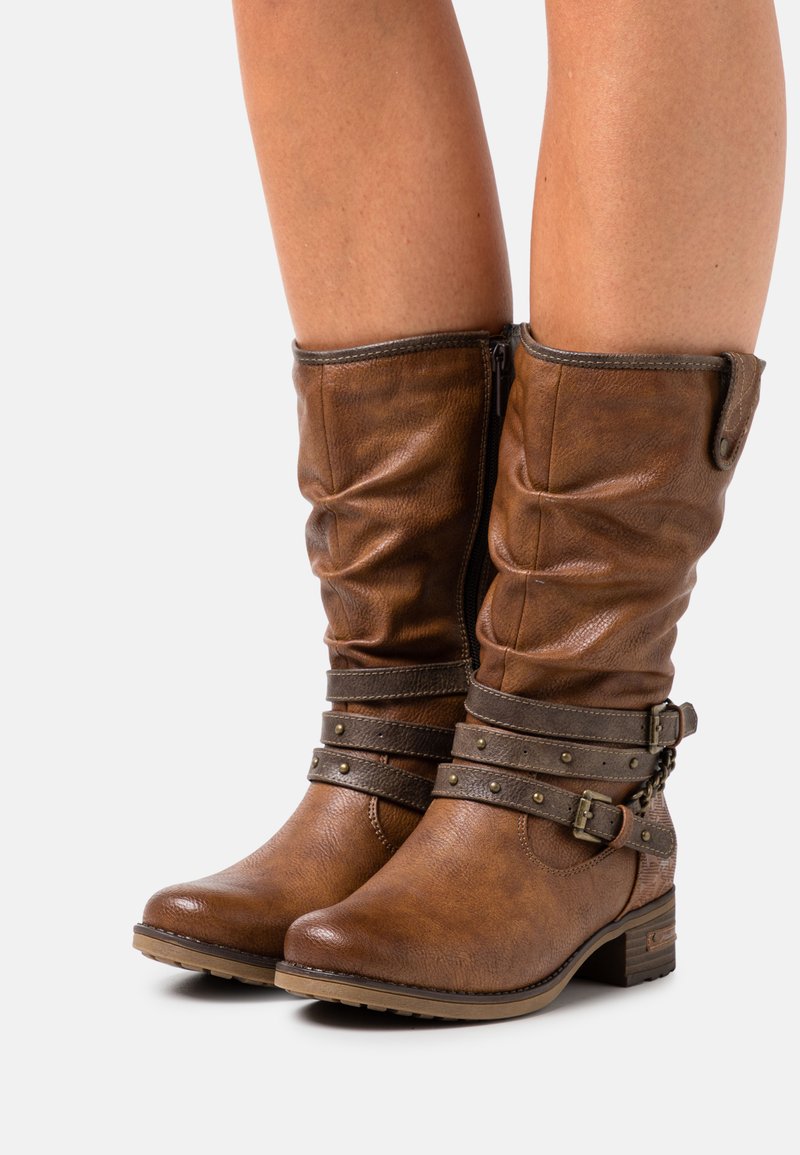 Bottes montantes en faux cuir marron avec tige ample, sangles décoratives, clous et fermeture éclair latérale. Talon bas épais pour plus de stabilité.