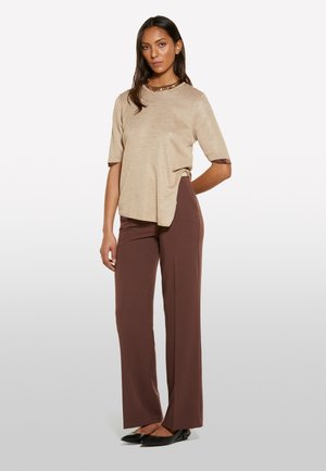 Donna con top beige a maniche corte, pantaloni marroni a gamba larga, ballerine nere e una collana d'oro massiccia, in piedi con una mano dietro la schiena.