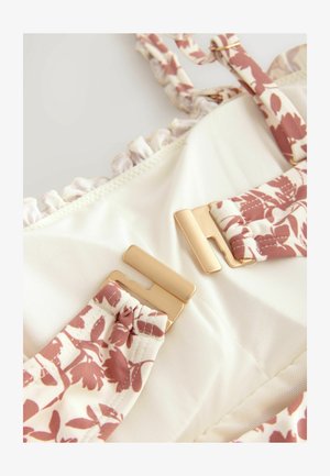 A cream-colored bikini top with floral pattern straps, gold hardware accents, and a frilled edge along the neckline.