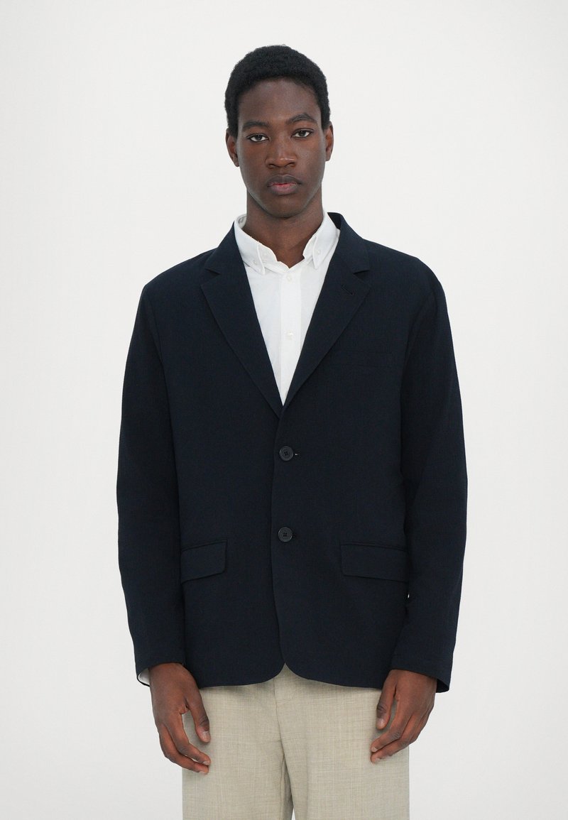 Navy blazer featuring two buttons, notch lapels, and front pockets, made of textured fabric. Paired with a white shirt and beige trousers.