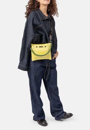 Frau trägt eine dunkelblaue, übergroße Jeansjacke und Hose mit schwarzen Schuhen, dazu eine leuchtend gelbe Umhängetasche mit geflochtenem Riemen.