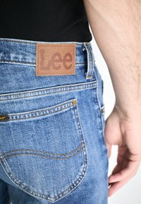 Blue denim jeans featuring a brown leather patch with "Lee" embossed. The design includes pockets, yellow stitching, and a slim fit.