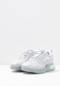 Ljusgrå träningsskor med en andningsbar mesh-övre, kurviga designdetaljer och genomskinlig luftdämpning på sulan. Röd logodetalj.