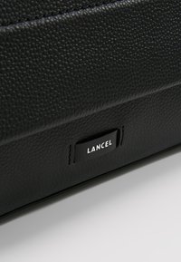 Cuir noir texturé avec un motif de grain subtil. Présente une forme rectangulaire et une petite étiquette rectangulaire embossée avec "LANCEL."