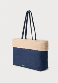 Geantă din denim cu o bordură din blană artificială bej, având două mânere de culoare albastru închis și o formă plată, structurată, cu un accent de logo auriu.