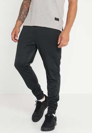 Mann trägt schwarze sportliche Jogginghose, schwarze Sneakers und ein hellgraues T-Shirt, steht mit einer Hand entspannt und der anderen leicht erhoben.