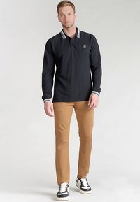 Zwarte polo met lange mouwen en witte streep aan de kraag, logo op de borst, gecombineerd met beige broek en zwarte sneakers met witte zolen.