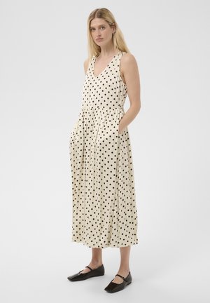 Femme aux cheveux blonds portant une robe crème sans manches à pois noirs et des chaussures plates noires sur un fond gris uni.